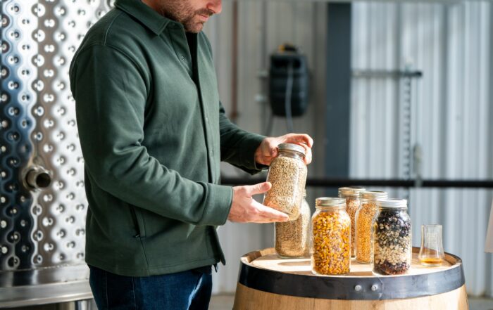 A distiller examines a mason jar filled with grain at West Fork Whiskey Co., surrounded by jars of assorted grains atop a whiskey barrel.