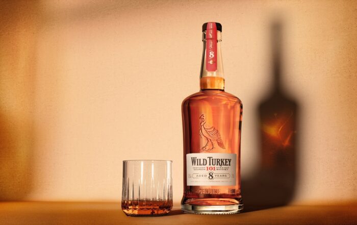 A bottle of Wild Turkey 101 8-Year-Old Bourbon next to a rocks glass half-filled with whiskey, set against a warm, softly lit background.